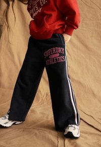Pantaloni della tuta navy con strisce laterali bianche e logo rosa "Superdry Athletics". Vestibilità ampia; texture morbida; abbinati a scarpe da ginnastica bianche.