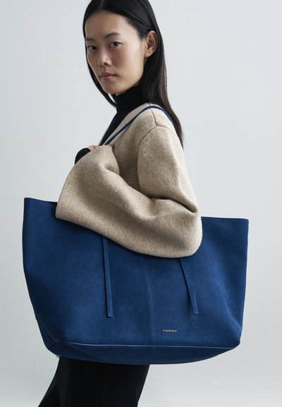 Blue suede tote bag with structured shape, featuring two long handles and subtle stitching details. Model wears a beige sweater.
