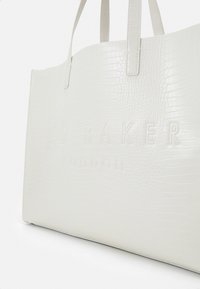 Bijela torba od krokodilne kože s utisnutim brandom "TED BAKER LONDON", koja ima strukturirani oblik i dva gornja ručka.