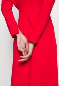 Largo vestido rojo con mangas largas, corsé ajustado y un detalle de cremallera en la muñeca, presenta manos entrelazadas detrás de la espalda.