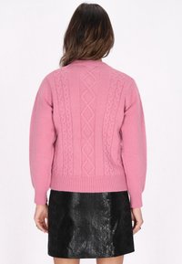 Rosa Kabelstrickpullover mit hohem Halsausschnitt, gerippte Bündchen und Saum. Kombiniert mit einem schwarzen, strukturierten Rock, der eine figurbetonte Silhouette zeigt.