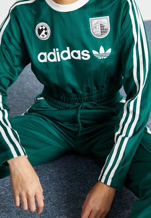Persoon draagt groene Adidas trainingspak met witte strepen en logo's, zittend op een blauw tapijt met handen die rusten nabij de knieën.