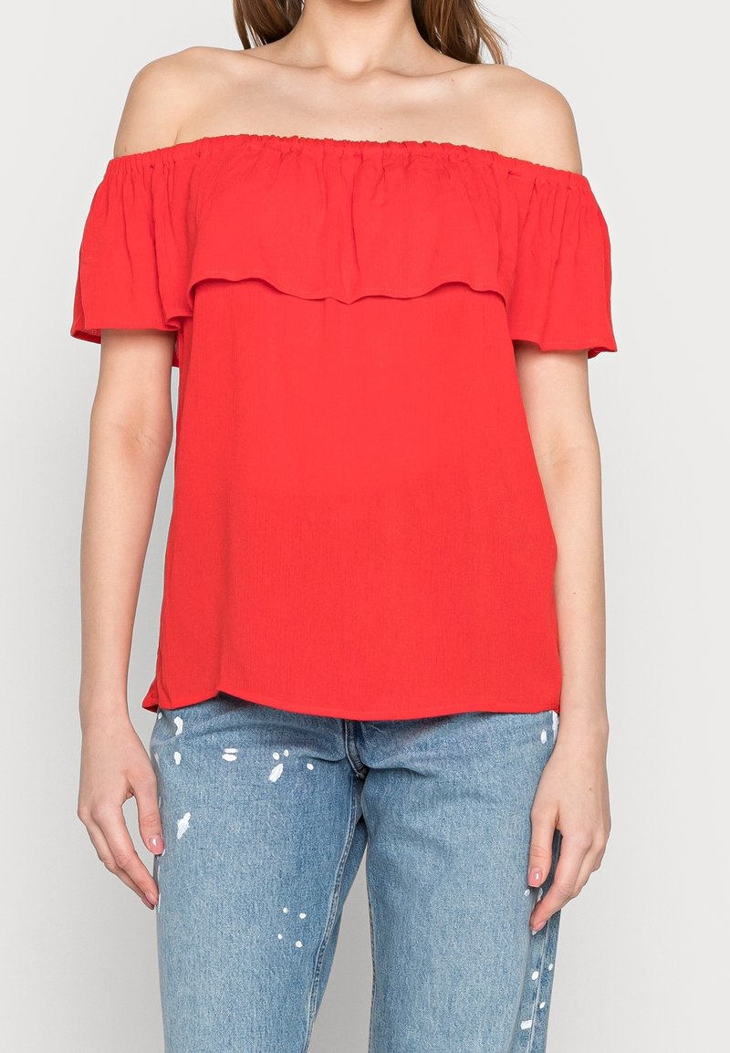 Top rosso off-the-shoulder con dettagli arricciati, realizzato in tessuto leggero. Abbinato a jeans in denim chiaro con dettagli di macchie di vernice.