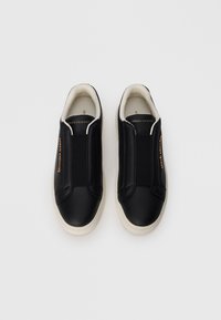 Sneakers neri slip-on con parte superiore in pelle liscia, pannelli laterali elastici e suola in gomma bianca testurizzata. Dettaglio del logo dorato sul lato.