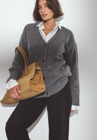 Grauer Strickcardigan mit V-Ausschnitt, ausgestattet mit großen Knöpfen. Kombiniert mit einem weißen Hemd mit Kragen und gehalten mit einer beigefarbenen Wildledertasche. Schwarze Hose sichtbar.