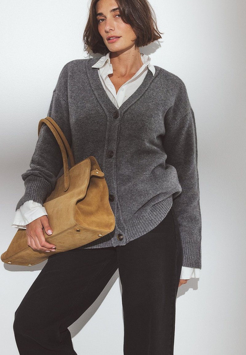 Grauer Strickcardigan mit V-Ausschnitt, ausgestattet mit großen Knöpfen. Kombiniert mit einem weißen Hemd mit Kragen und gehalten mit einer beigefarbenen Wildledertasche. Schwarze Hose sichtbar.