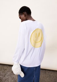 Camicia bianca a maniche lunghe con un grande volto sorridente giallo ricamato sulla schiena, abbinata a una gonna di jeans e che tiene un cappellino bianco.