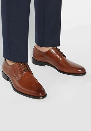 Brown leather dress shoes with a polished finish, pointed toe, and six eyelets laced with thin, matching brown laces, paired with navy trousers.