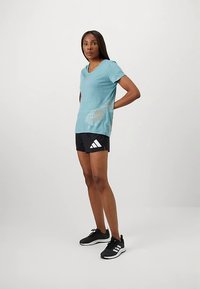 Kobieta stoi z rękami za plecami, ubrana w jasnobłękitny T-shirt z kwiatowym wzorem, czarne szorty Adidas i czarne trampki na białym tle.