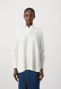 Camicia bianca con colletto, maniche lunghe e vestibilità morbida, abbinata a una gonna di jeans con uno spacco frontale. Tessuto dalla texture liscia.