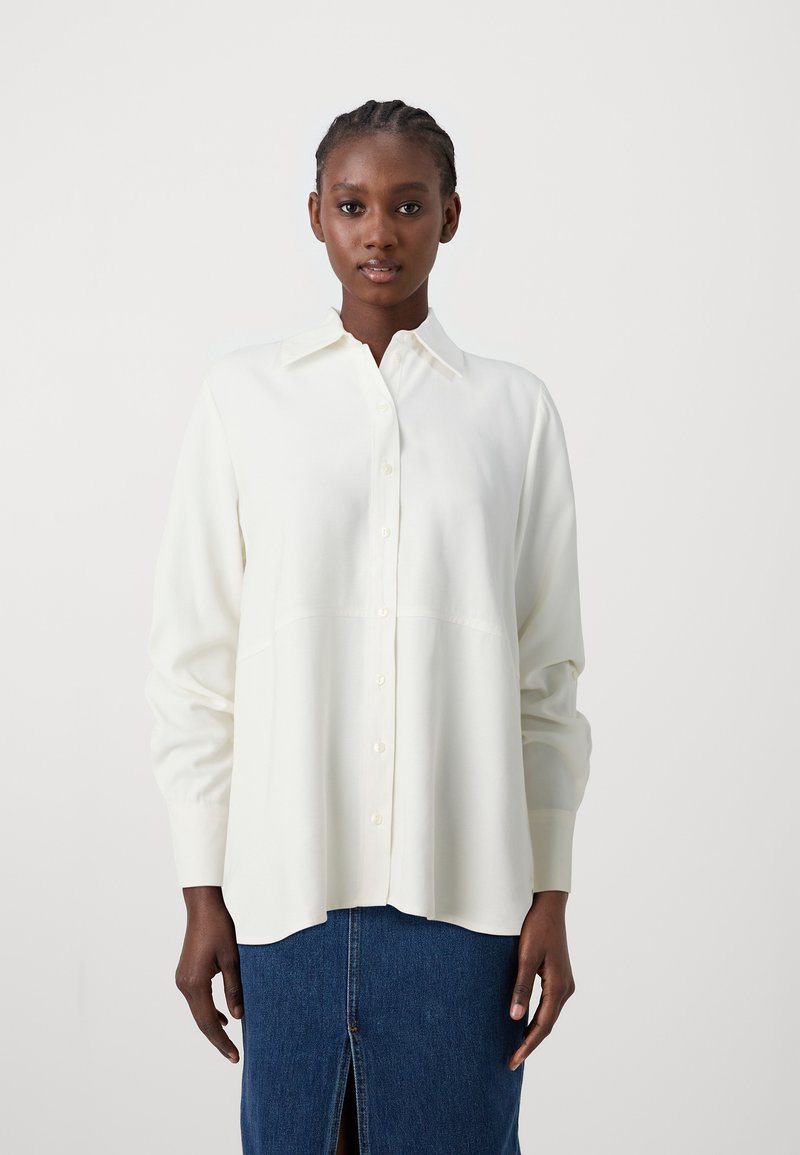 Camicia bianca con colletto, maniche lunghe e vestibilità morbida, abbinata a una gonna di jeans con uno spacco frontale. Tessuto dalla texture liscia.