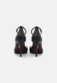Czarne szpilki z błyszczącej skóry, z wąskim czubkiem i smukłym obcasem stiletto. Posiadają regulowany pasek na kostce.