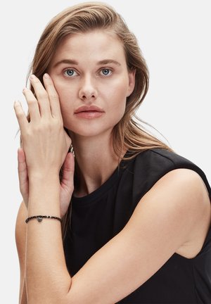 Schwarzes Perlenarmband mit einem kleinen Herzakzent, am Handgelenk getragen, vor einem schlichten Hintergrund. Das Modell hat lange Haare und einen neutralen Ausdruck.