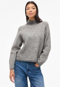 Suéter cinza de malha com um padrão de espinha de peixe, gola alta e corte solto; combinado com jeans de ganga azuis. Textura suave e estilo descontraído.