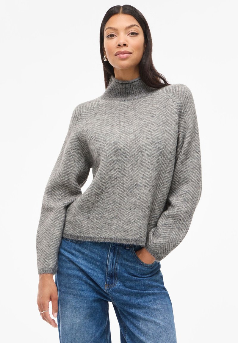 Suéter cinza de malha com um padrão de espinha de peixe, gola alta e corte solto; combinado com jeans de ganga azuis. Textura suave e estilo descontraído.