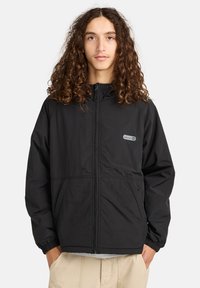 Junge Person mit langen lockigen Haaren, die eine schwarze Jacke mit Reißverschluss und Fronttaschen sowie beigefarbene Hosen trägt, stehend mit Händen in den Taschen.