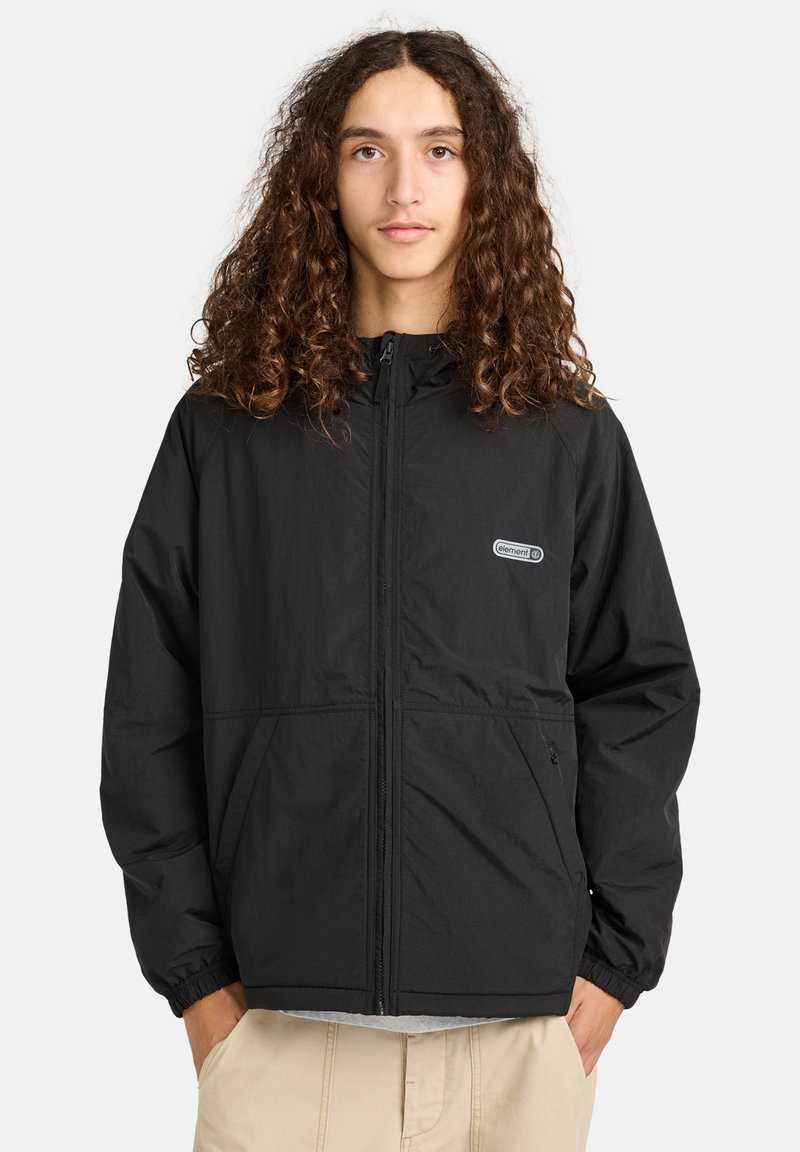 Junge Person mit langen lockigen Haaren, die eine schwarze Jacke mit Reißverschluss und Fronttaschen sowie beigefarbene Hosen trägt, stehend mit Händen in den Taschen.
