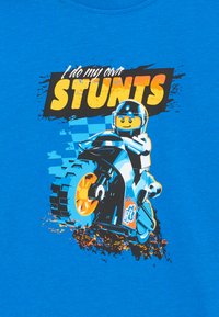 Blaues T-Shirt mit einem Grafikdesign eines Cartoon-Figuren auf einem Motorrad mit orangefarbenen Rädern, daneben der Text "Ich mache meine eigenen STUNTS" in fettem Gelb.
