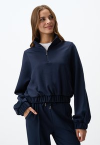 Maglione pullover blu navy con colletto a mezza zip, vita elastica e polsini arricciati. Tessuto morbido e design rilassato adatto per un abbigliamento informale.
