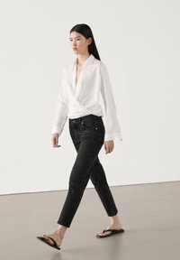 Femme marchant portant un chemisier blanc style cache-cœur, un jean noir slim et des tongs noires sur un sol lisse et neutre.