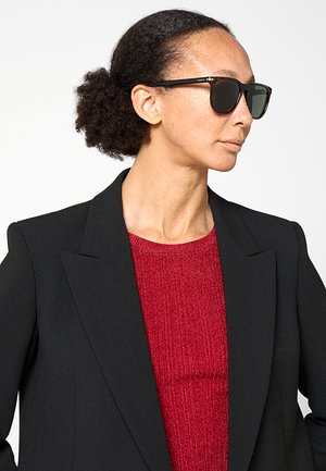Donna con capelli scuri e ricci raccolti, occhiali da sole neri, blazer nero e top rosso strutturato, rivolta verso destra su sfondo bianco.