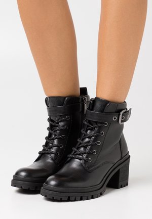 Černé kožené kotníkové boty s robustním podpatkem, vybavené šněrováním, prošívaným límcem a bočním zipem. Obsahují dekorativní pásek s přezkou.