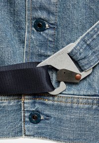 Hebilla de metal unida a una correa azul marino sobre denim azul claro. Incluye características como costuras visibles y una etiqueta metálica con marca.