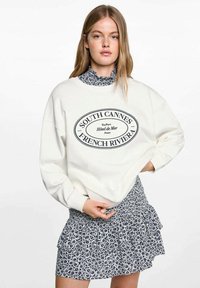 Valkoinen huppari, jossa musta soikea graafinen teksti "SOUTH CANNES FRENCH RIVIERA." Yhdistettynä tummansiniseen kukkakuosisesti röyhelöskriin.