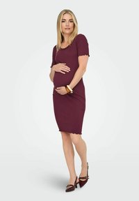 Femme enceinte portant une robe moulante bordeaux et des talons assortis, caressant doucement son ventre, debout devant un fond clair uni.