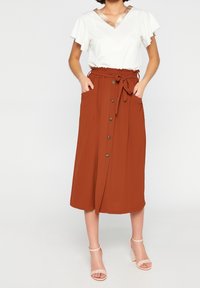 Gonna midi color ruggine con bottoni frontali, tasche laterali e vita annodata. Abbinata a una maglietta bianca con scollo a V e collo decorato.