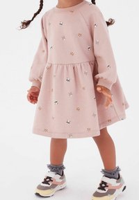 Ein Kind trägt ein pinkes Kleid mit Tiermotiven, das gepuffte Ärmel und einen ausgestellten Rock hat, kombiniert mit bunten Turnschuhen und grauen Socken.