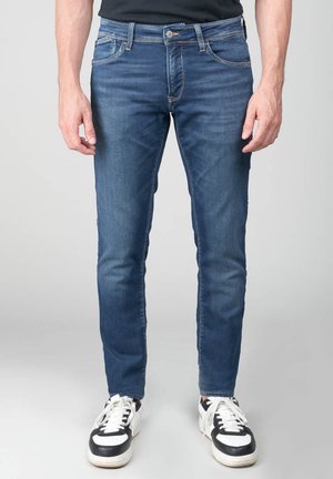 Vaqueros de mezclilla azul oscuro con corte ajustado, con cinco bolsillos, sutil desgaste y cremallera. Se llevan con zapatillas negras y blancas.