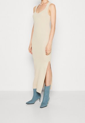 Kvinde iført en ærmeløs beige ribstrikket midi-kjole med en sideslids og blå ruskindede støvler med hæl, stående mod en hvid baggrund.