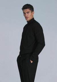 Veste zippée noire au design ajusté, fabriquée en tissu tricoté texturé, avec un col haut et une silhouette élancée, associée à un pantalon noir.