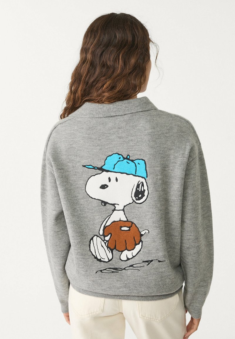 Suéter gris de punto con diseño de cuello. Presenta un gráfico colorido de Snoopy en la espalda, que lleva una gorra azul y una camisa naranja.