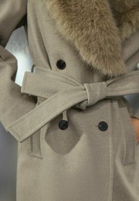 Beige wollen jas met een nep-fur kraag, dubbelknopen ontwerp, zwarte knopen en een ceintuur in de taille, met een gladde textuur en gestructureerde vorm.