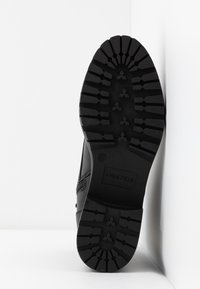 Semelle de botte noire avec des motifs de bande de roulement profonds et le nom de la marque "Anna Field" embossé près du talon sur un fond blanc.