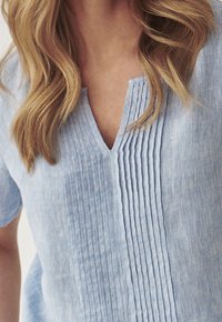 Blonde vrouw in een lichtblauwe linnen blouse met verticale plooien en een kleine V-halsopening.