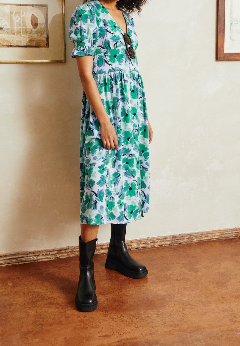 Blommigt klänning i blå och gröna nyanser, med puffärmar och v-ringning, kombinerad med svarta ankelstövlar och en strukturerad beige golv.