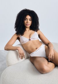 Gaiši rozā apakšveļas komplekts ar trīsstūra formas krūšturi un sarkanu un zilu logotipu, kopā ar baltām biksītēm ar logotipa jostiņu.