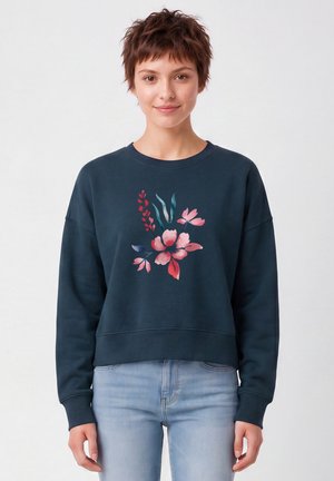 Jeune femme aux cheveux courts et bruns porte un sweat-shirt bleu foncé décoré d’un imprimé floral rose et rouge, associé à un jean bleu clair.