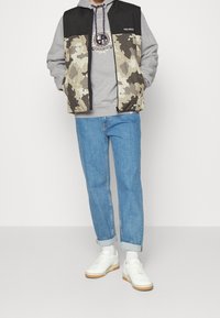 Chaleco acolchado con patrón de camuflaje en beige y negro, combinado con una sudadera gris, jeans azules y zapatillas blancas con suelas de goma.