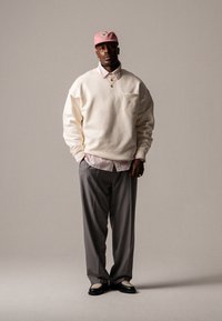 Kremfarget sweatshirt med lomme, lys rosa skjorte med krage, grå bukser og svarte loafers. Rosa caps med logo. Enkel, avslappet passform.