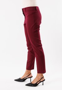 Pantaloni scuri in borgogna su misura con una vestibilità slim e lunghezza alla caviglia, dotati di tasche laterali e una texture liscia, abbinati a décolleté nere con cinturino.