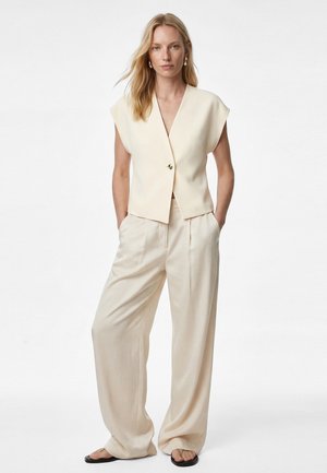 Mujer con cabello largo rubio, vestida con top crema sin mangas abotonado, pantalones crema de pierna ancha, pendientes de aro dorados y sandalias negras, con las manos en los bolsillos.