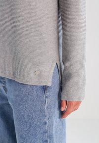 Helles graues Sweatshirt mit horizontalen Streifen, gerippten Bündchen, seitlichen Schlitzen und einem kleinen Logo-Akzent am Saum. Kombiniert mit blauen Jeans.