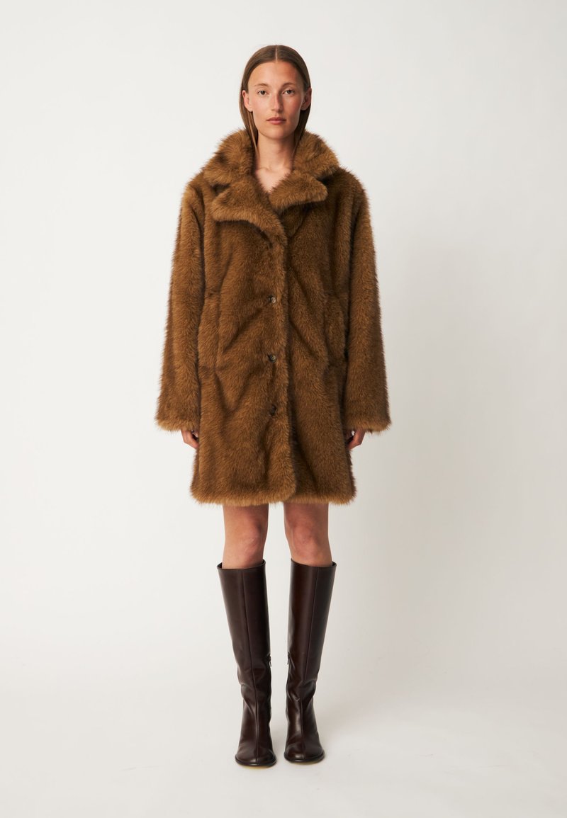 Femme debout portant un manteau mi-long en fausse fourrure marron avec des boutons et des bottes en cuir foncé montant jusqu'aux genoux sur un fond clair uni.
