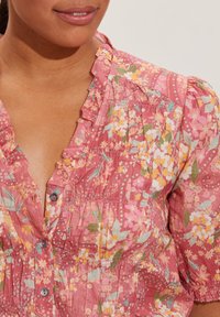 Pink floral blouse with a ruffled neckline, short sleeves, and button detail. Features layered pleats and a mix of colorful flower patterns.
