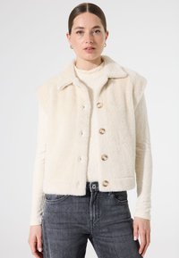 Kunstbont mouwloze vest in crème met een kraaghalslijn, vijf grote ronde knopen en textuurstof. Gecombineerd met grijze jeans.