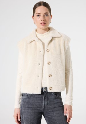 Gilet sans manches en fausse fourrure crème avec un col, cinq grands boutons ronds et un tissu texturé. Associé à un jean gris.
